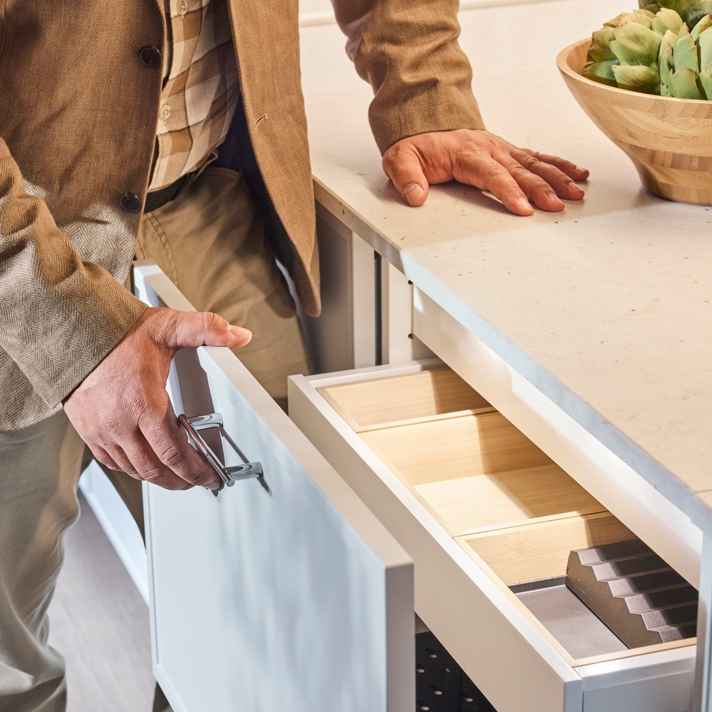 Hand opens kitchen drawer with wooden organizers while other rests on countertop; bowl of greens sits nearby in modern space.