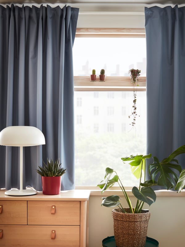 Cortinas BENGTA celestes a medio cerrar filtrando la luz de una habitación. Delante de la ventana hay una cómoda BJÖRKSNÄS con una lámpara de mesa NYMÅNE.