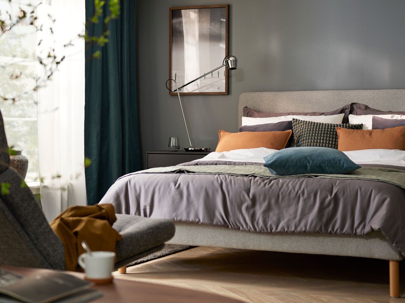 A bedroom with grey walls and view of bed, bedside table and shelves. Blurry green leaves from a plant in the foreground.