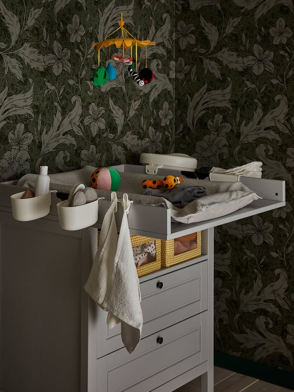Une table à langer équipée d’un matelas à langer SKÖTSAM avec housse, des accessoires, des jouets et un mobile suspendu au-dessus.