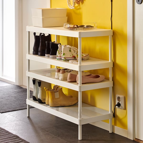 In a hallway with a bright yellow wall is a white MACKAPÄR shoe rack with four shelves filled with shoes in various styles.