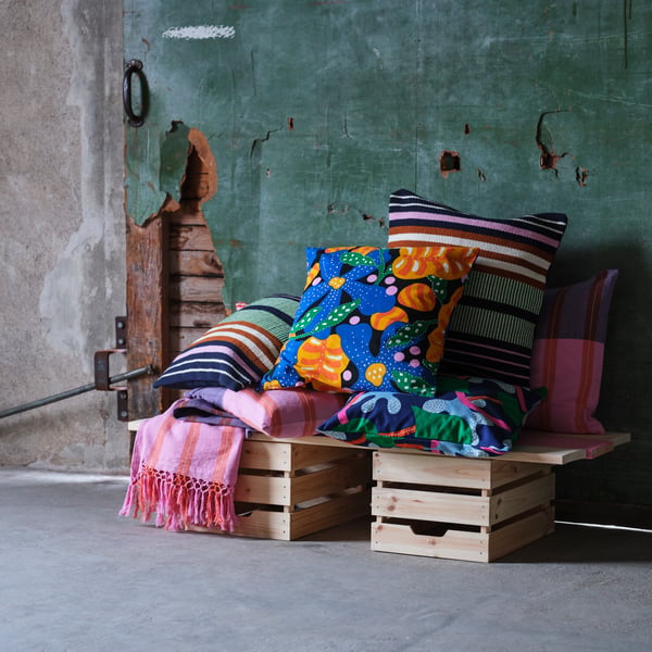 Sur deux boîtes en bois, des housses de coussin et des jetés de la collection MÄVINN en rose, bleu, vert et orange.