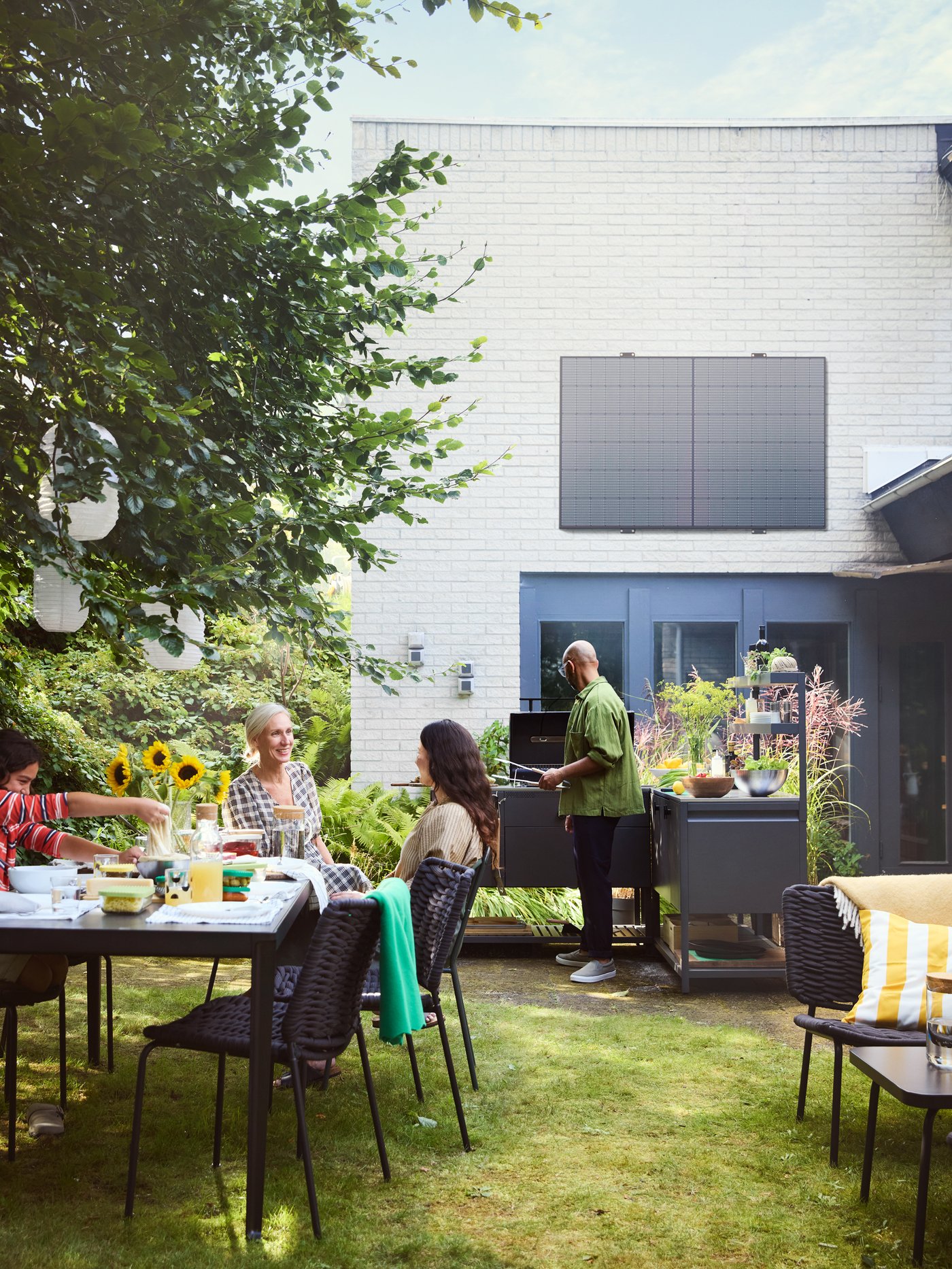 Un groupe d'amis profite d'un rassemblement estival en plein air, une personne faisant des grillades sur une cuisine extérieure IKEA GRILLSKÄR tandis que d'autres sont assis autour d'une table à manger IKEA avec des chaises VASSHOLMEN, sous un arbre feuillu orné de lanternes en papier blanches.