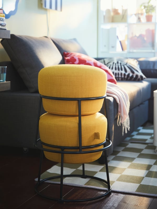 Two yellow EMMISLÖV pouffes, one stacked on top of the other, stand on the edge of a white/green KLASSRUM rug near a sofa.