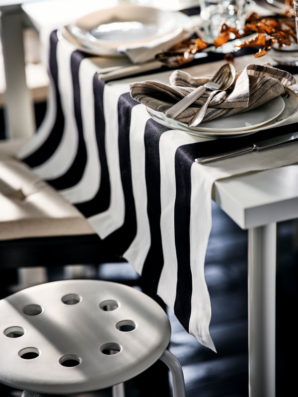 A corner of a decked-out table with white OFTAST plates and beige UPPHÖJD cutlery. Beneath stands a white MARIUS stool.