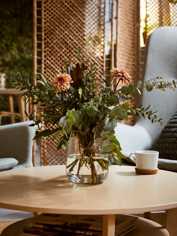 A leafy flower bouquet in a BERÄKNA clear-glass vase on a JAKOBSFORS coffee table in a softly sunlit lounge area.
