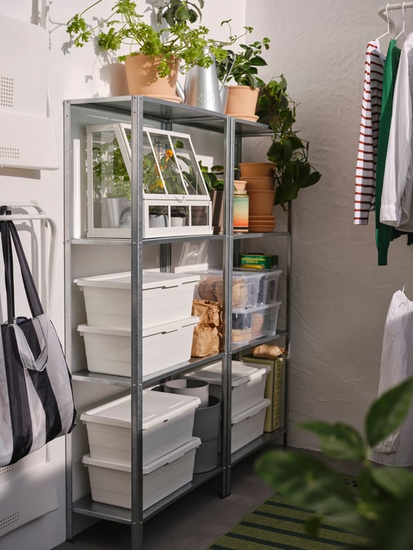 Two HYLLIS shelving units with stacked boxes with lids, potted plants and a white greenhouse, plus a carrier bag on a hook.