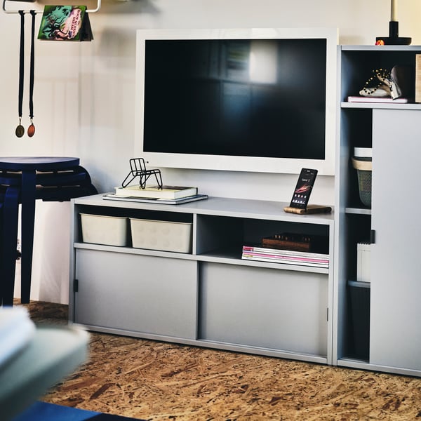 Téléviseur au cadre blanc fixé au mur au-dessus d’un banc TV SPIKSMED gris clair, à côté d’un rangement SPIKSMED gris clair.