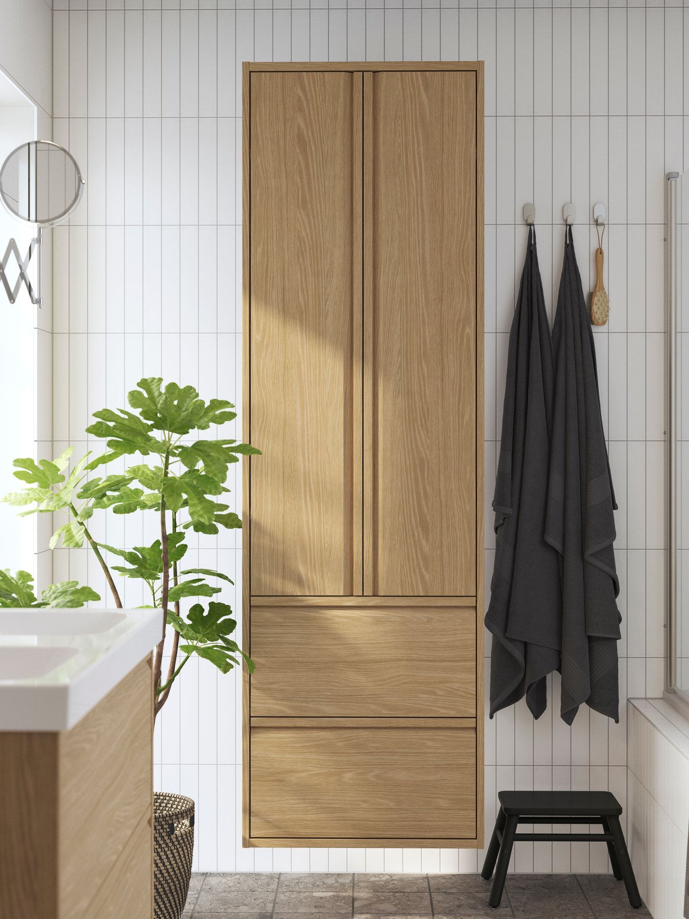 Une grande armoire en bois IKEA est fixée sur un mur de salle de bains carrelé en blanc, à côté de serviettes sombres suspendues à des crochets, d’une plante verte, d’un miroir rond et d’un tabouret noir.