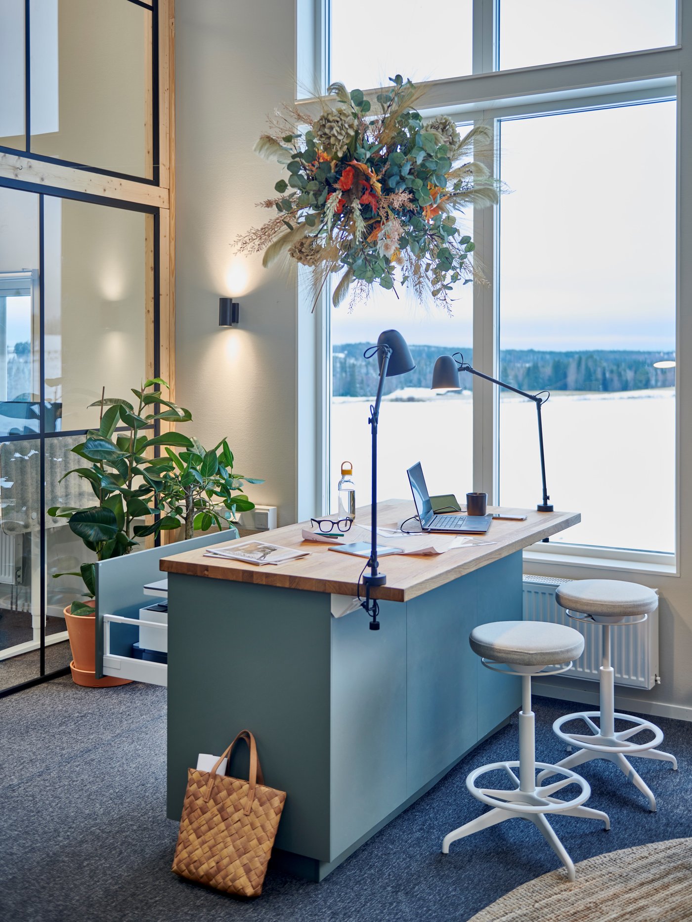 Modernt kontor med stort fönster, skrivbord, två vita pallar, lampor, växter och en hängande blomdekoration ovanför.