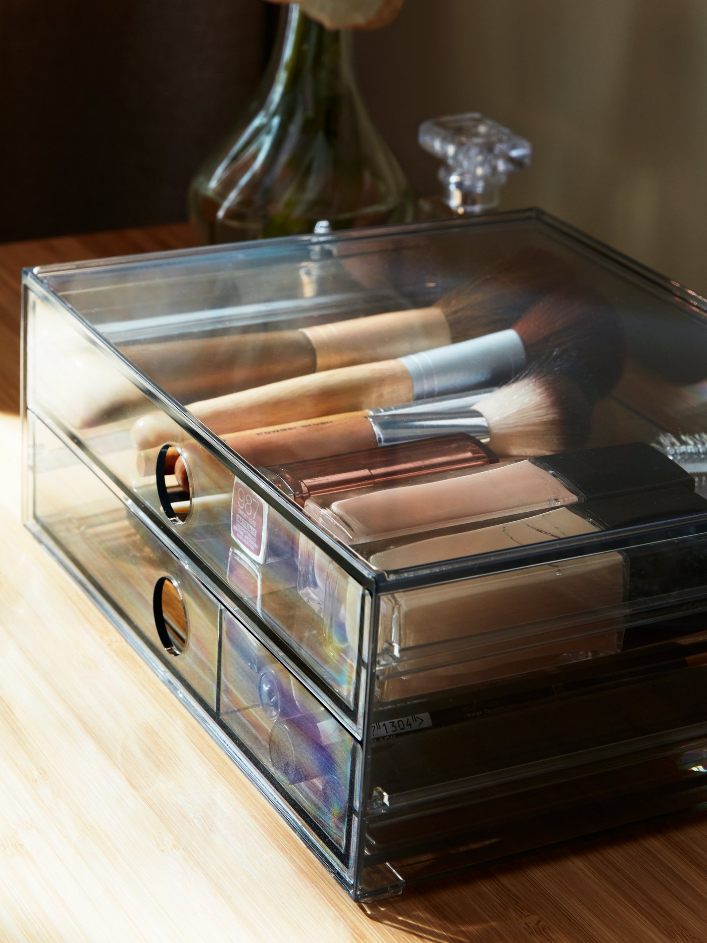 Organisierte Make-up-Produkte sind in einer rauchfarbenen GODMORGON Minikommode mit 2 Schubladen neben einer Blumenvase auf einem Frisiertisch zu sehen.