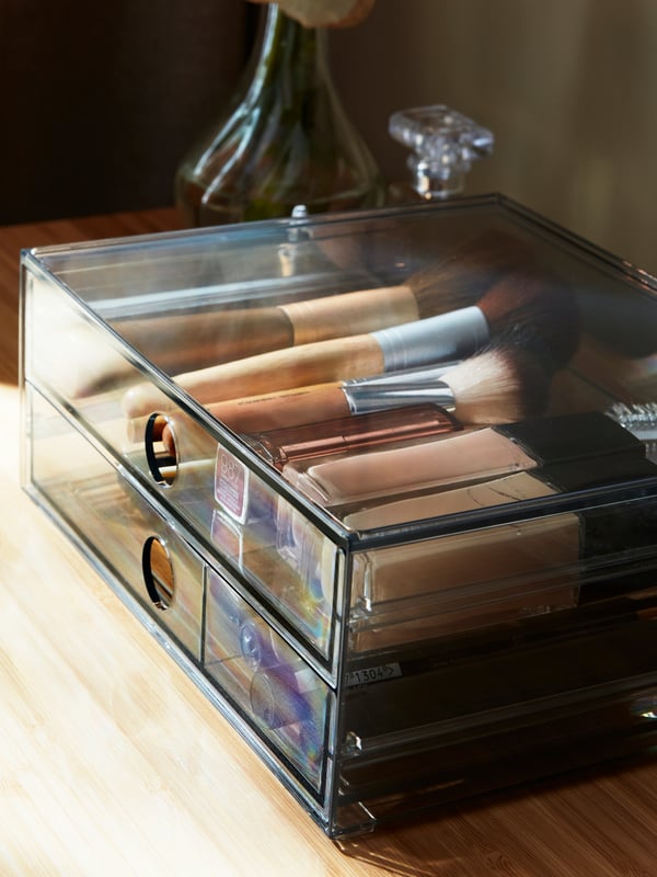 Organised makeup products in a smoked GODMORGON mini chest with 2 drawers next to a flower vase on a dressing table.
