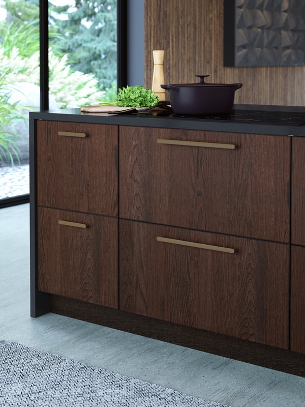 A kitchen island with wooden drawers and bronze handles, a chopping board with greens on it, and a black cooking pot.