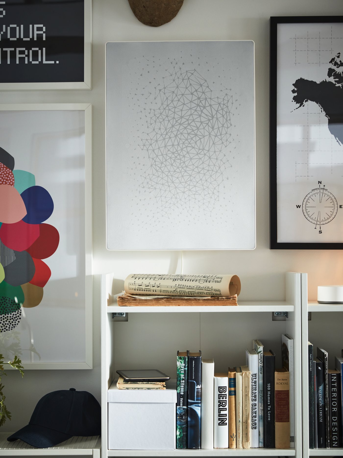 Framed art and a white picture frame with WiFi speaker on the wall, white bookcases, books, a cap, a sheet of music.