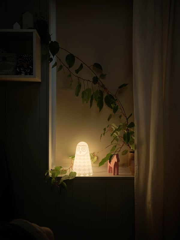 Prozor sa zatvorenom FÖNSTERBLAD roletnom za zamračivanje na čijem simsu je bela SOLBO LED stona lampa u obliku sove.