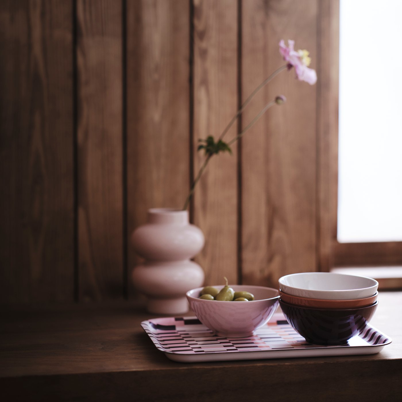 A close-up of HÖSTAGILLE bowls in pink, brown, off-white and orange on a patterned tray by a pink vase on a windowsill.