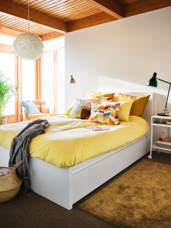 A bedroom with a white MALM bed frame, yellow quilt covers and cushions, and a HANNELISE cushion with a colourful pattern.