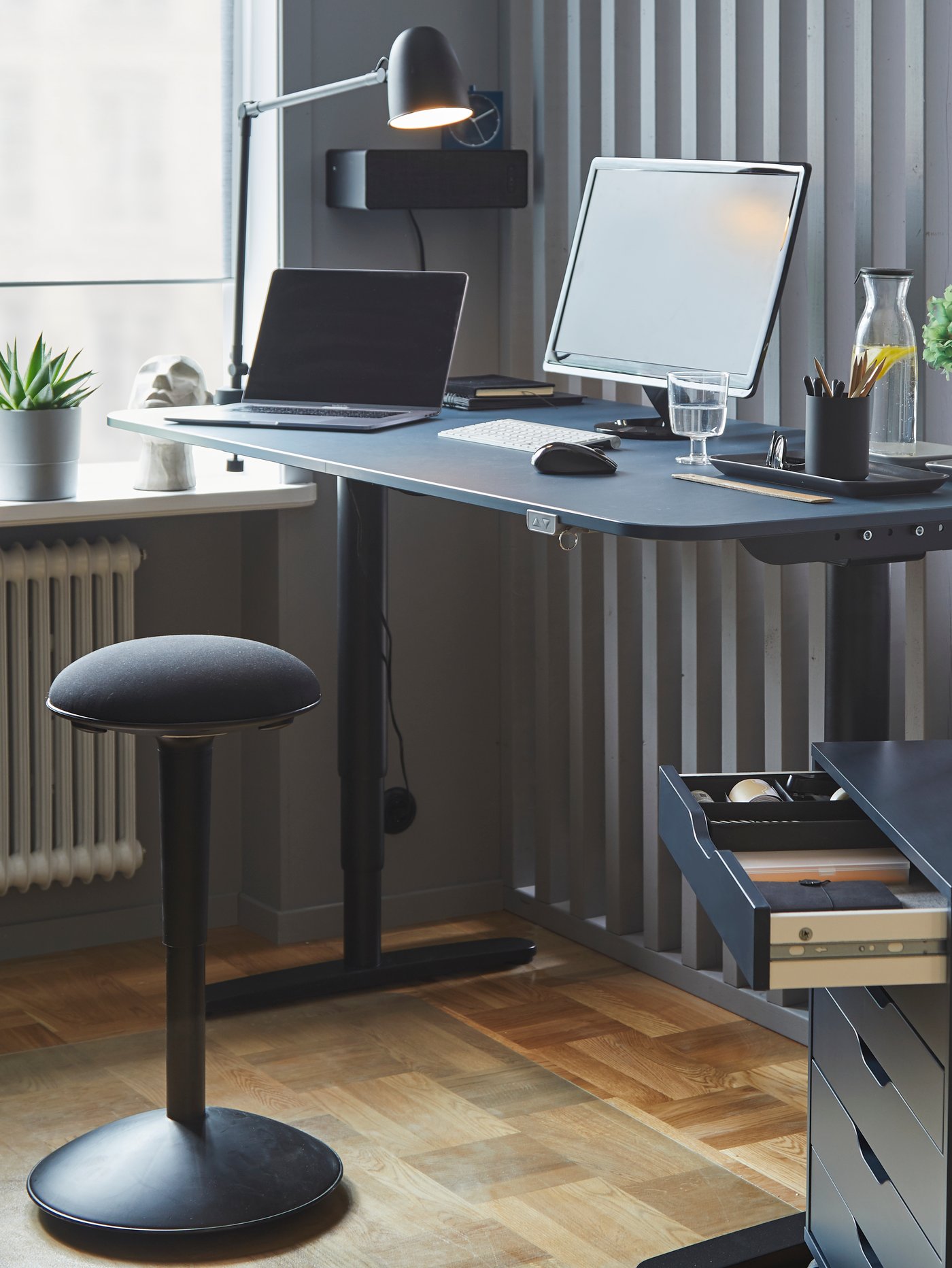 Uno sgabello NILSERIK nero/Vissle nero davanti a una scrivania per seduta attiva con uno schermo da computer in uno spazio di lavoro - IKEA