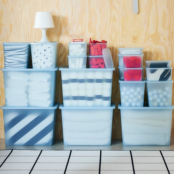 Small to large RYKTA storage boxes in translucent blue colour stacked neatly on top of each other against a wood grain wall. 