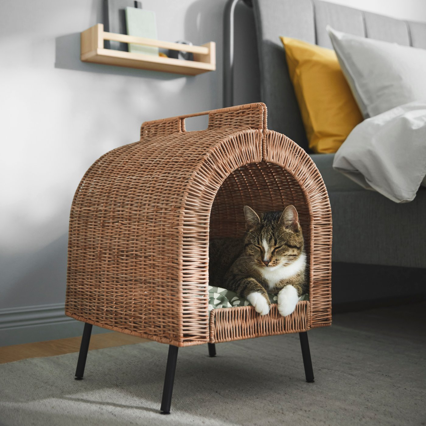 A tabby-and-white cat rests inside a raised wicker-style cat house with an arched entrance and black metal legs. The cat house sits in a modern bedroom beside a gray upholstered bed with white bedding and a mustard-yellow accent pillow. A small wooden wall shelf is mounted on the light gray wall, and soft natural light creates a warm, cozy atmosphere.