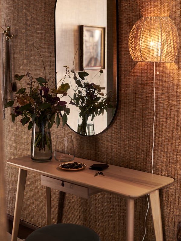 A black LINDBYN mirror and a lit MÅNALG lamp hanging side-by-side on a wall above an ash-veneer LISABO desk with decorations.