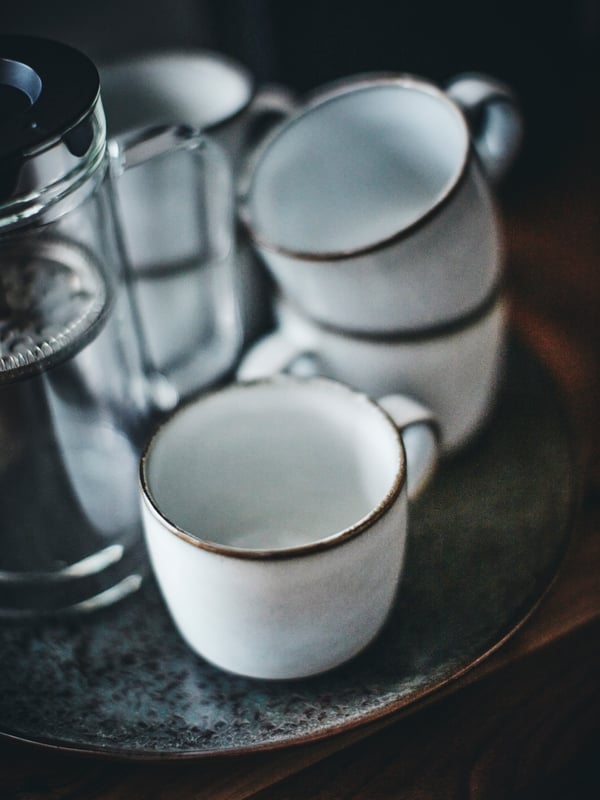 Des tasses GLADELIG grises sont empilées les unes sur les autres sur une assiette GLADELIG à côté d’une cafetière.