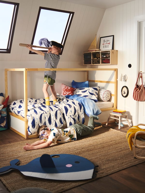 A boy stands on a bed dressed in a BLÅVINGAD whale duvet cover pretending to be a pirate; another boy plays on the floor.
