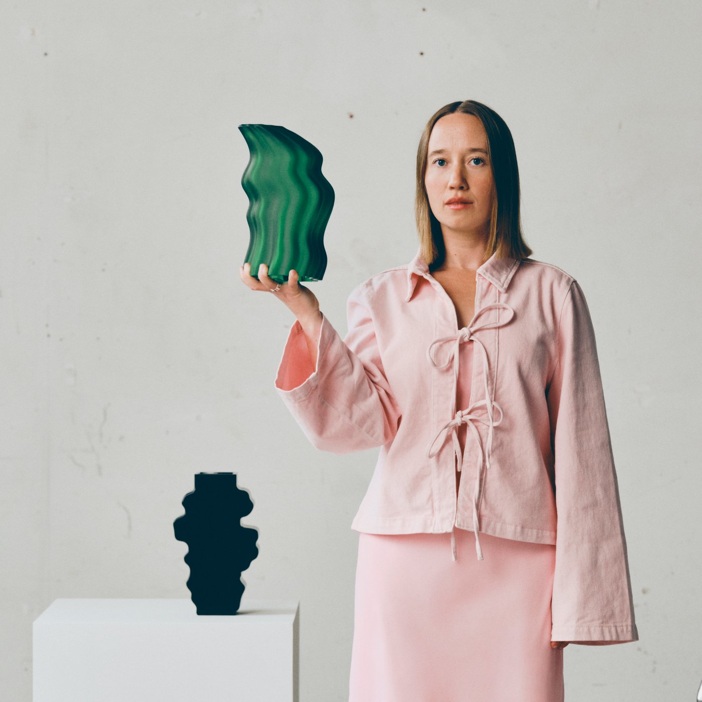 Person in pink clothing holds a green wavy vase beside a black decorative sculpture on a white pedestal.