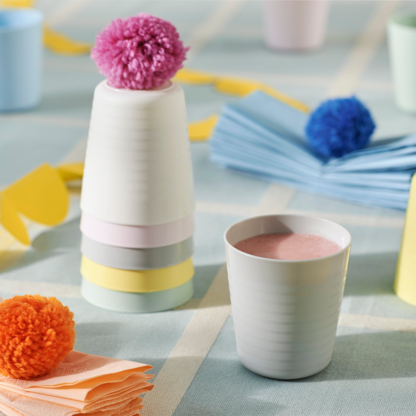 Various pastel colour KALAS children’s mugs on a light blue check table cloth. One is filled with a pink smoothie.