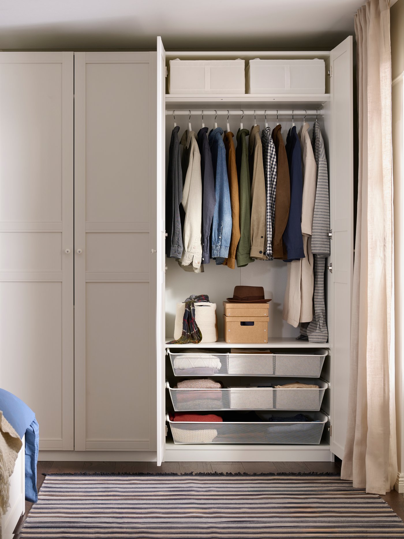 A bedroom with an open white PAX wardrobe with clothes hanging inside and KOMPLEMENT mesh baskets at the bottom.