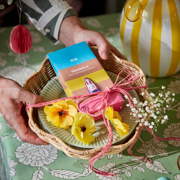 Personne tenant une corbeille à pain contenant un sachet de lapins en chocolat et des fleurs artificielles.