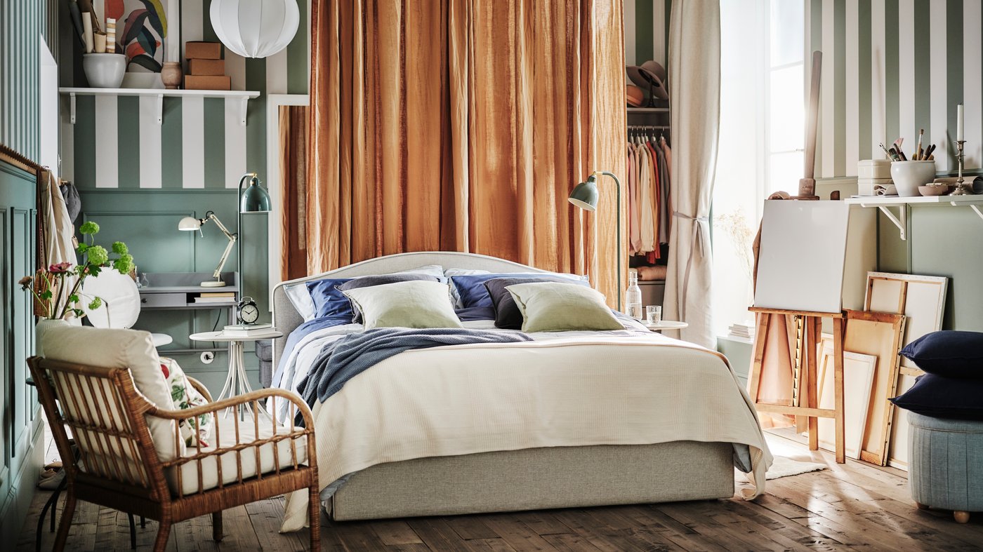 A beige RAMNEFJÄLL upholstered bed in a green and brown room by a HOLMSTA armchair with dark beige DYTÅG curtains behind it.