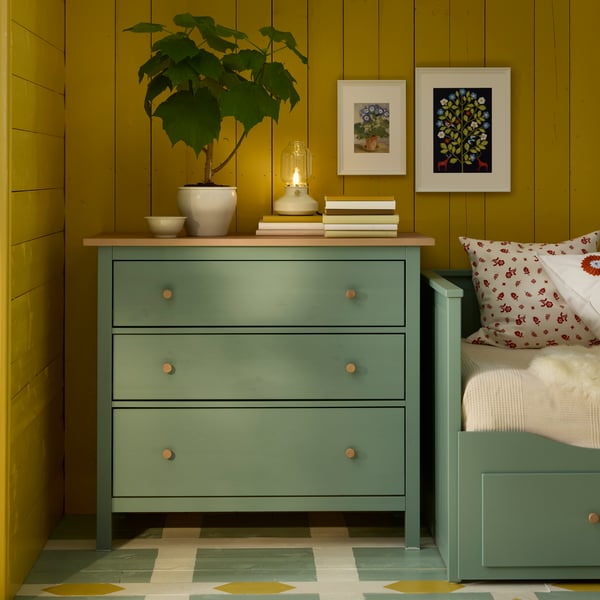 옐로 컬러의 벽을 배경으로 한 공간에 그레이그린 HEMNES 헴네스 3칸 서랍장 위에 TÄRNABY 테르나뷔 탁상스탠드, 화분이 놓여 있는 모습.