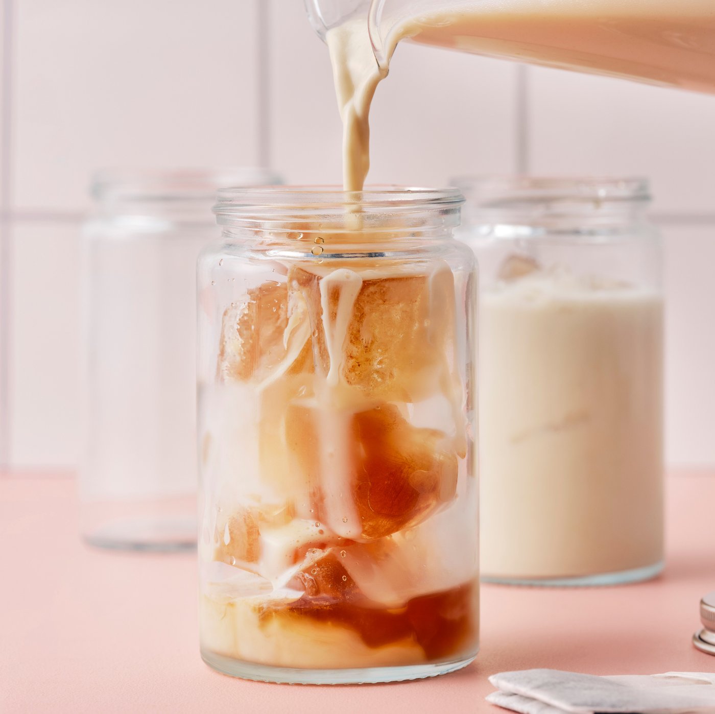 Eine Person gießt Haferdrink für Kaffee aus einer Kanne in ein Glas, das mit Eiswürfeln aus Wasser und FLÖJT Schwarztee gefüllt ist.