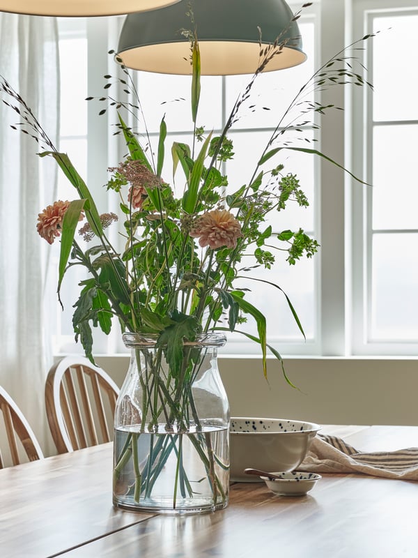 A clear glass BEGÄRLIG vase with flowers in it stands on a SKOGSTA dining table near some windows in a dining room.