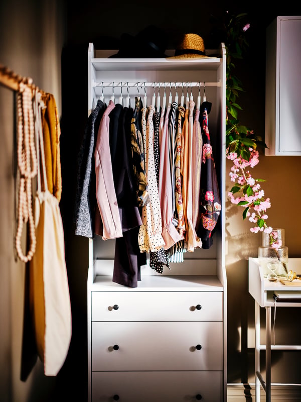 Ein weißer HAUGA offener Kleiderschrank mit 3 Schubladen. An einer Kleiderstange hängt Kleidung, auf einem Schreibtisch zur Rechten steht eine Vase mit Blumen.