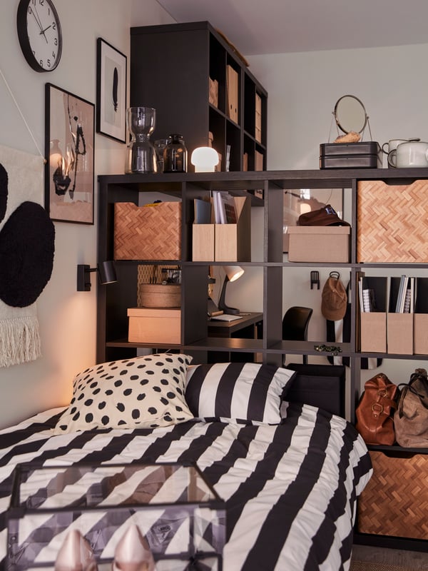 A compact dorm room with a large, black-brown KALLAX shelving unit with inserts and boxes by the head end of a bed.