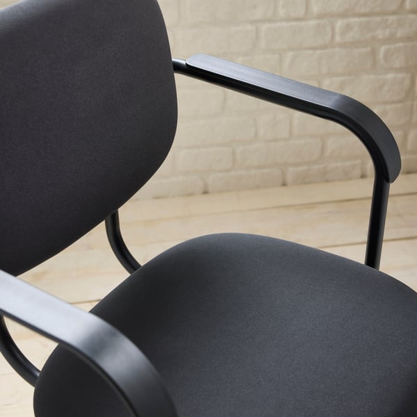 A close-up of a dark grey MULLSJÖ swivel chair against a white brick wall, standing on a wooden floor.