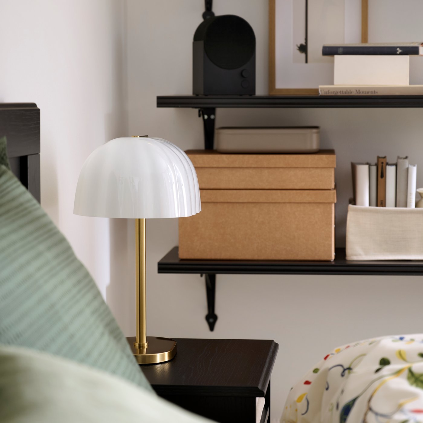 The corner of a bedroom with AKTERSPRING table lamp beside the bed and black shelves behind.