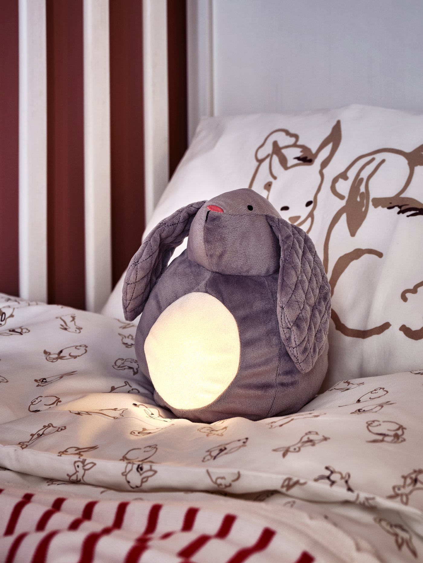 Mainan lembut dengan lampu malam LED PEKHULT bentuk arnab terletak di atas katil bayi SUNDVIK berwarna putih dengan linen katil RÖDHAKE.