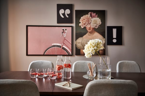 Konferensrum med ett mörkt träbord och  grå stolar. På bordet står två karaffer med vatten, glas och en vas med vita blommor. På väggen bakom syns tavlor med citattecken, ett porträtt av en kvinna med blommor över ansiktet, ett utropstecken och ett rosa cykelhjul.