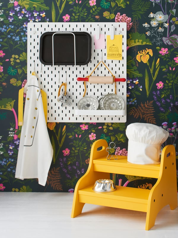 Kitchen accessories such as an apron and a rolling pin are hung on a white SKÅDIS Pegboard. A chef's hat is placed on a yellow TROGEN Children's step stool, which is placed in front of the pegboard.