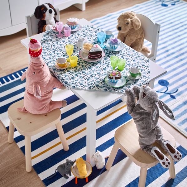Cuatro peluches con formas de animales sentados alrededor de una mesa infantil preparada para el té con accesorios de juguete. Están sentados sobre pisos FLISAT y sillas infantiles SUNDVIK, que se encuentran sobre una alfombra con rayas azules y blancas. 