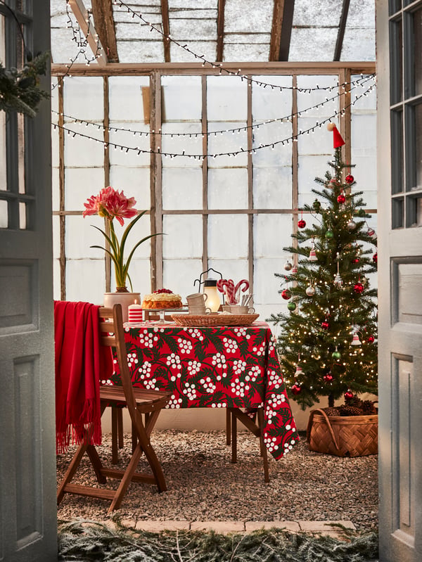 Afternoon tea / fika in greenhouse at Christmas time with red and white VINTERFINT tablecloth, artificial tree and LED lights.