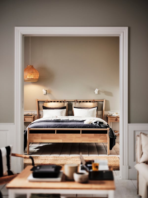 Ein modernes Schlafzimmer mit einem Holzrahmenbett, weißen Kissen und einer gemütlichen Decke. Dezente Wandleuchten und ein Naturfaserteppich sorgen für eine warme, einladende Atmosphäre. Minimalistisches Design, perfekt für jedes Zuhause.