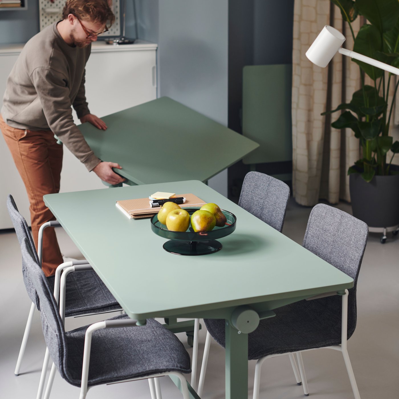 A small meeting area with two green MITTZON foldable table with castors, four grey chairs and a man moving one table.