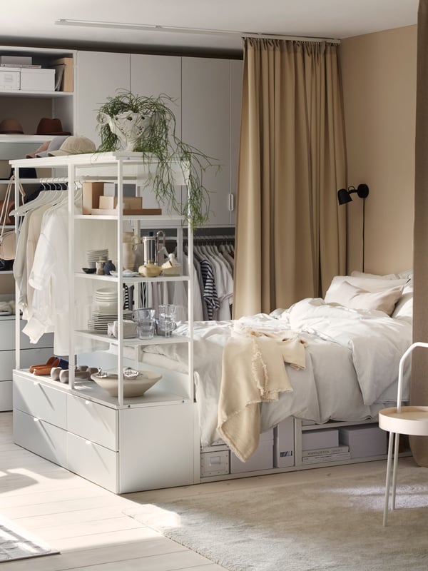 Rangka katil dengan empat laci PLATSA berwarna putih, peralatan tempat tidur berwarna putih di atas katil, pakaian tergantung pada rangka, terdapat barangan pada para-para.
