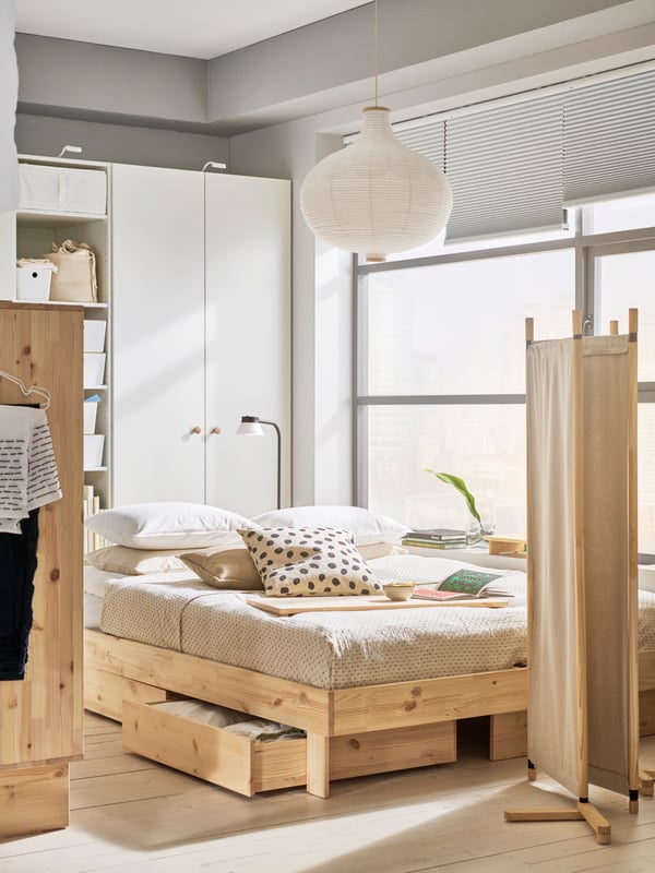 Bedroom in beige colours with GLAMBERGET bed and room divider in natural wood. White wardrobe and big window in back.