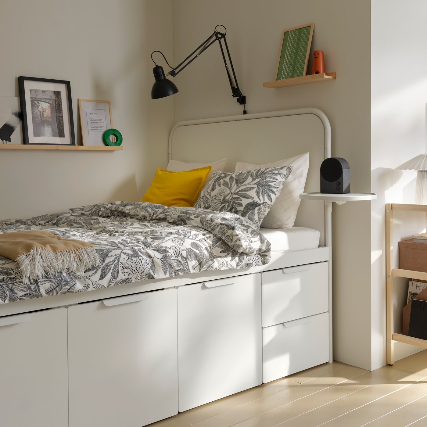 In una stanza per studenti in affitto, una struttura letto con contenitori MÅSSKÄR bianca con un piumino a fiori bianco e nero contro una parete angolare.