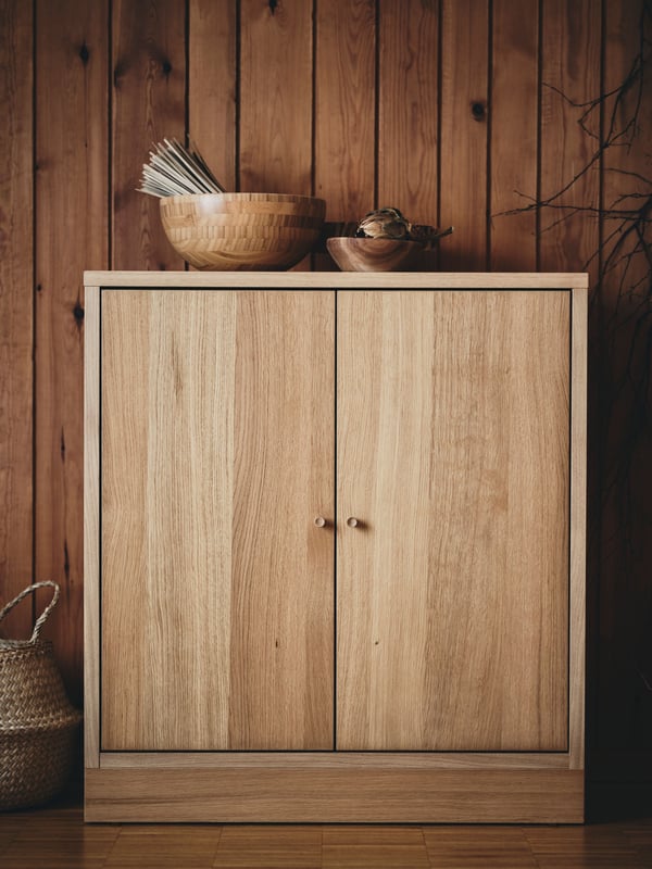 Nahaufnahme eines TONSTAD Schranks mit Türen aus Eichenfurnier, der vor einer holzvertäfelten Wand steht. Obenauf sind zwei Holzschüsseln platziert.